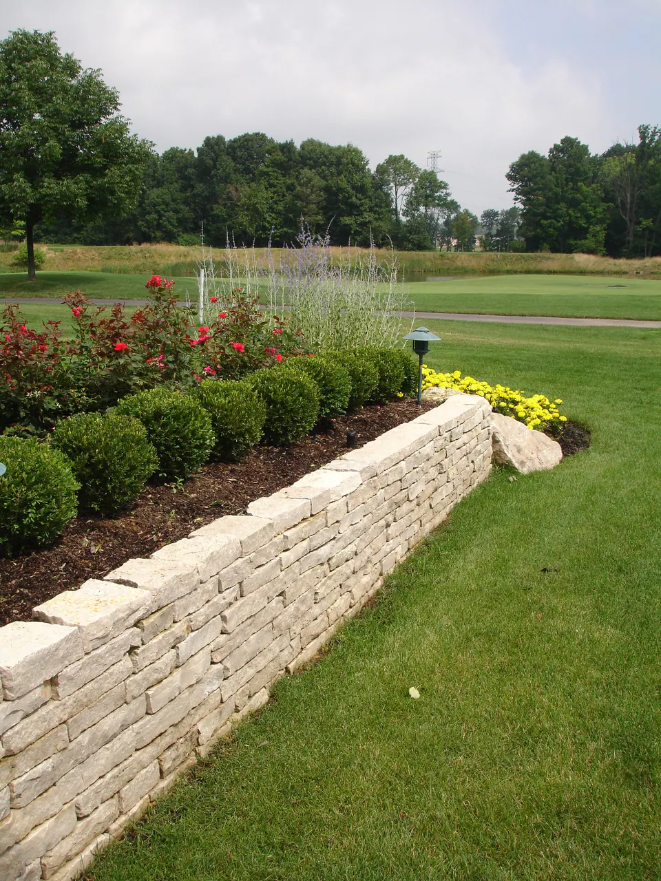 Low dry-laid limestone wall holding a planting bed of boxwood and roses beside a lawn